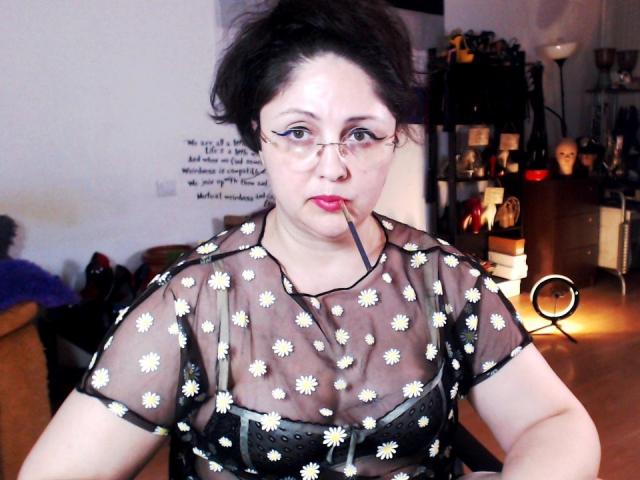 Photograph of ImperatrizaSanda with pale skin, red lipstick, and black hair, wearing a sheer black top with white daisy patterns, exposing her cleavage, glasses, and a suggestive look. Background is blurred.
