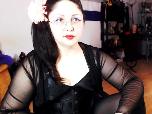 Fotografie von ImperatrizaSanda, einer Frau mit heller Haut und schwarzen Haaren im Pferdeschwanz, die ein durchsichtiges schwarzes Oberteil trägt und korsett, roter Lippenstift, helles, weißes Licht im Gesicht, dunkler Hintergrund, sexuelle Spannung..