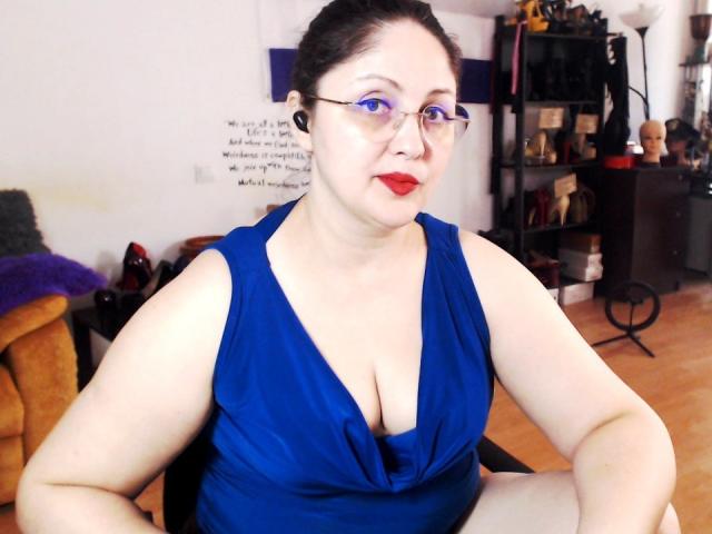 ImperatrizaSanda, plus-sized with pale skin, black hair in a bun, wearing red lipstick and glasses, deep blue sleeveless dress with cleavage, seated, black shelves with objects in the background.