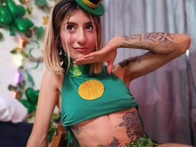 Kaiix, con i capelli biondi, indossa un top verde a vita alta con un cerchio giallo, ha tatuaggi sulle braccia e sull'addome, è appoggiata con una mano, in posizione leggermente inclinata. sorriso, fascia verde per capelli, sfondo festivo, posa sensuale, atmosfera informale.