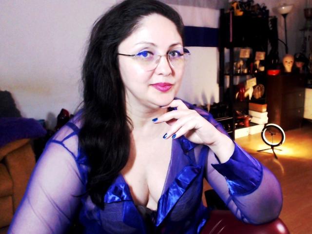 ImperatrizaSanda, con piel pálida, cabello negro y gafas, vestida con una blusa azul transparente que deja ver parte de su escote. Ella está posando con un dedo en los labios, irradiando tensión sexual. Iluminación brillante.