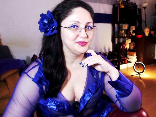 Imperatriza Sanda, mit heller Haut und schwarzen Haaren, trägt ein durchsichtiges blaues Kleid mit einer blauen Rose im Haar und sitzt eine Hand in der Nähe ihres Gesichts, leicht verführerischer Ausdruck. Helle Beleuchtung.