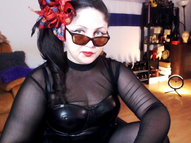 Imperatriza Sanda, con piel pálida y cabello negro, vestida con una blusa negra transparente sobre un corpiño de cuero negro, y flores rojas en el cabello. accesorio, gafas de sol marrones. El fondo muestra muebles y luces desenfocadas. Tensión sexual..