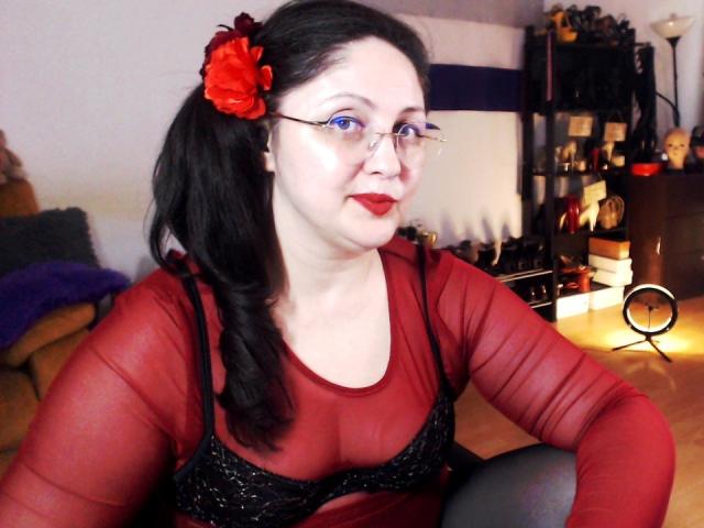 ImperatrizaSanda mit heller Haut und schwarzen Haaren, trägt rote Brillen und ein durchsichtiges rotes Oberteil mit einem schwarzen BH, eine rote Blume. in ihren Haaren, sitzend, sexy, verführerisch, zur Seite blickend.