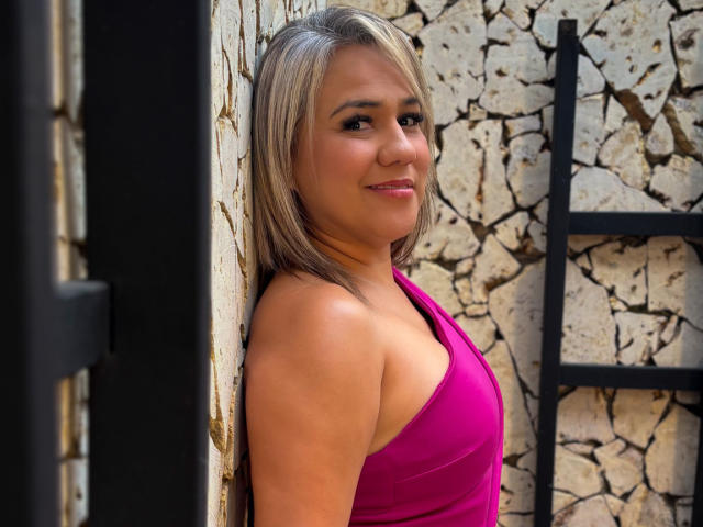 Fotografia de CamilaNoer encostada em uma parede de pedra, vestindo uma regata rosa justa, sorrindo com olhos castanhos, cabelos lisos. cabelo loiro, pele clara, atraente, decote discreto, ambiente externo.