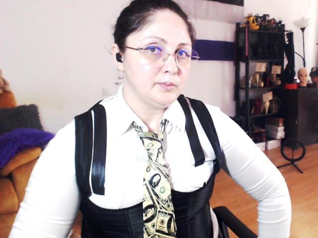 Fotografie von ImperatrizaSanda mit heller Haut, schwarzen Haaren zu einem Dutt gebunden, die eine Brille trägt, ein weißes Hemd, ein schwarzes Korsett und krawatte mit Muster, sitzend in einem Wohnzimmer, leicht unscharf, intensiver Blick..