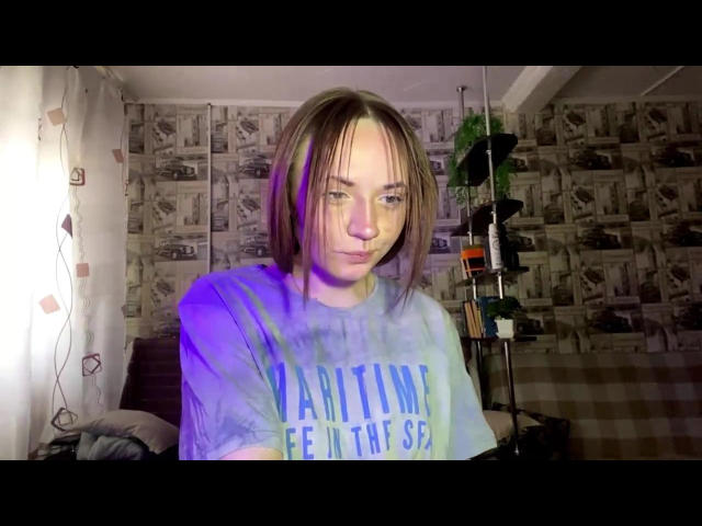 RonnieWeeda, mit schulterlangem braunem Haar, trägt ein blaues T-Shirt mit der Aufschrift "ICH BIN KEIN KÄMPFER" und sitzt in einem Raum mit Wänden. Poster, die von sanftem Licht erhellt werden und eine stimmungsvolle, leicht erotische Atmosphäre schaffen..