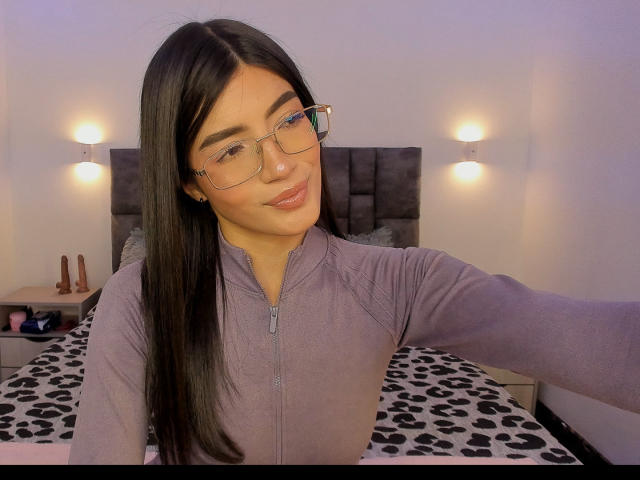 PaulethJoens, con lunghi capelli neri, che indossa occhiali e una felpa con zip viola, seduta su un letto con un motivo a pois bianchi e neri. fogli con puntini, illuminazione soffusa, sorriso appena accennato, sexy, ambientazione da camera da letto.