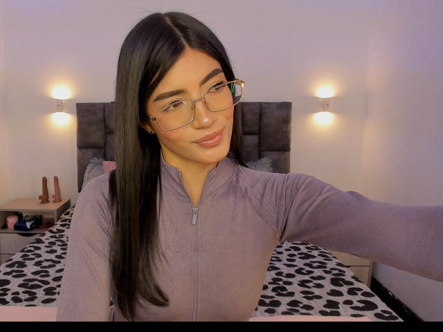 PaulethJoens, wearing a purple zip-up hoodie, glasses, and a slight smile, takes a selfie in a bedroom with black-and-white patterned bedspread, soft lighting, and two wall-mounted lamps. Fucking sexy.