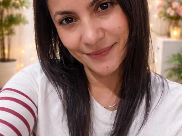 Close-up photo of SolaraDubois, a woman with long black hair, brown eyes, and a slight smile, wearing a white shirt with red stripes. She has a cute mole under her right eye. Fucking attractive.
