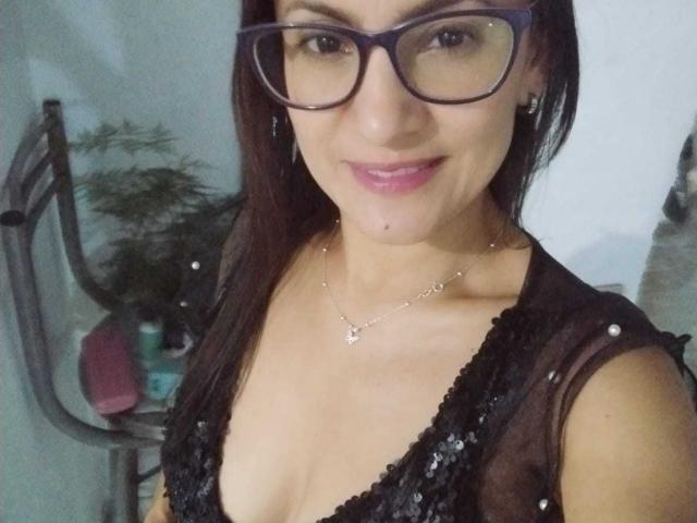 SolaraDubois mit schwarzen Brillengläsern, lächelnd, trägt ein durchsichtiges schwarzes Kleid mit Pailletten, das tiefe Ausschnitte zeigt, und eine Halskette. mit Kreuzanhänger, dunkles Haar, Innenraum, subtile sexuelle Spannung, lockeres Selfie.