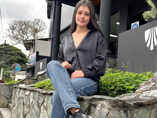 LizzaRose with long dark hair, sitting on a stone ledge, wearing a black button-up shirt and blue jeans, legs crossed, black sneakers, sexy, slightly blushed, outdoor setting, modern architecture.