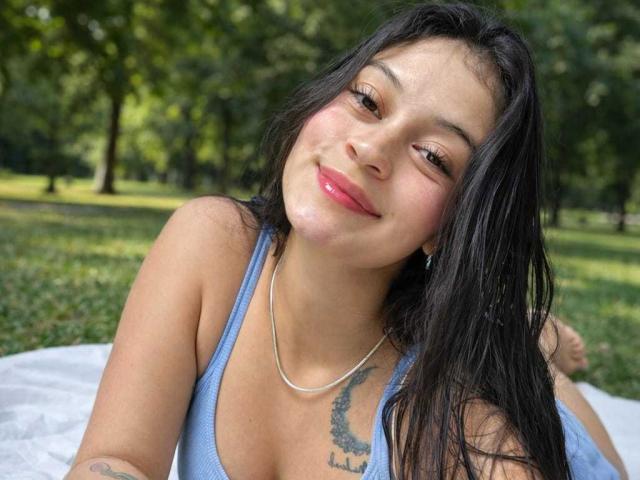 Primer plano de LizzaRose con cabello largo y negro, acostada sobre una manta blanca en un parque. Ella lleva una camiseta sin mangas azul, está sonriendo y tiene un tatuaje en el lado derecho de su pecho. Qué lindo/a..