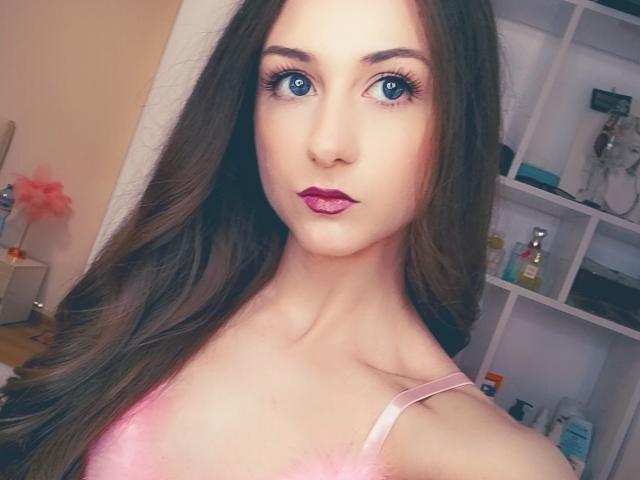 Close-up of Bessarida with large blue eyes, dark brown hair, and pink lipstick. She wears a pink top, looking directly at the camera with a slight, sexy smirk. Shelf with toiletries in the background.