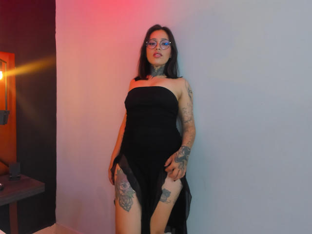 PaisleyMour stands against a white wall, wearing a black strapless dress, revealing tattooed thighs. She has dark hair, glasses, and a sexy, confident look with red and blue lighting.