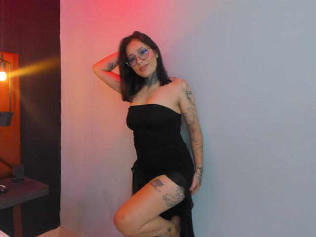 PaisleyMour leans against a wall, wearing a strapless black dress, exposing tattoos on her arm and thigh. She has glasses, black hair, and a sexy, confident pose. Red and blue light highlights her.