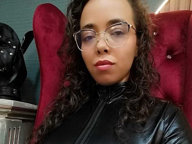 Barbara Leythonn, con gafas y una chaqueta de cuero negro, está sentada en una silla de terciopelo rojo, mirando directamente a la cámara. con la boca ligeramente abierta, transmitiendo un aire sexy y seguro..