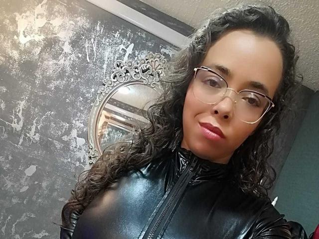 Foto van Barbara Leythonn met krullend zwart haar, die een bril draagt en een zwarte leren jas aanheeft, en die recht in de camera kijkt. camera met een subtiele glimlach. Achtergrond: gestructureerde muur, ronde spiegel. Ongelooflijk aantrekkelijk..