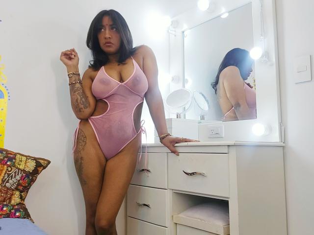 Nahirr, una mujer latina de curvas, con cabello oscuro, vestida con lencería rosa de una sola pieza con detalles recortados que realzan sus grandes senos. y tatuajes. De pie frente a un espejo de tocador brillante, luciendo seductora..