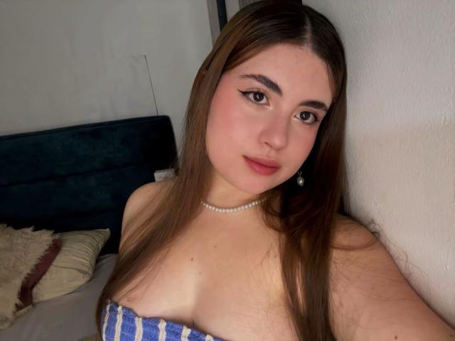 VicluX met lang bruin haar, lichte huid, die een blauw-witte gestreepte top zonder bandjes draagt, een parelketting, en die naar iets kijkt. camera, lichte glimlach, zacht licht, bed op de achtergrond, seksuele spanning.