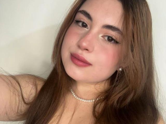 Close-up of VicluX with long brown hair, light skin, and brown eyes, wearing a pearl necklace, looking directly at the camera with a slight smile. Foreground focus on face. Sexual tension present.