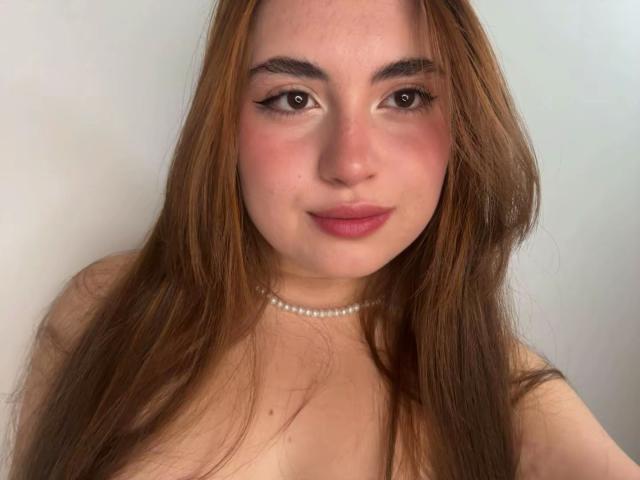 Gros plan de VicluX, avec de longs cheveux bruns, portant un collier de perles et sans haut, laissant entrevoir une légère décolleté. Sourire doux, maquillage léger, lèvres roses. Fond neutre. Légère tension sexuelle..