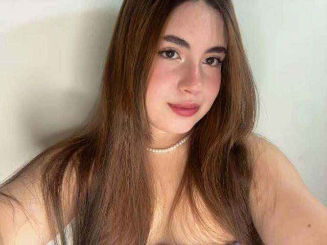 Photograph of VicluX, a fair-skinned woman with long brown hair, wearing a pearl necklace, topless, smiling slightly, light pink lipstick, soft shadow on the right side, subtle sexual tension.