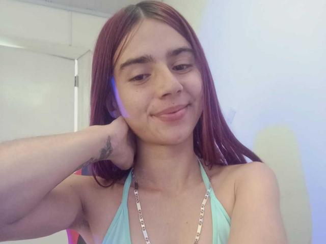 Fotografia de uma jovem mulher, LoveMaxim, com cabelos castanhos longos, sorrindo levemente, vestindo uma blusa cropped azul clara com colar de corrente. Ela tem uma tatuagem no braço esquerdo. Sua expressão é relaxada, com um toque de tensão sexual..