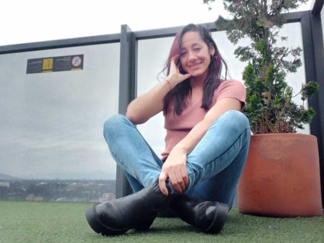 SamanthaCorine, a brunette woman with a medium build, sits cross-legged on green turf, wearing a pink t-shirt and blue jeans, smiling with her hand on her cheek. Black boots, potted plant.