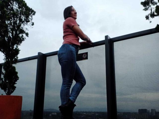 SamanthaCorine staat op een balkon, leunend tegen een glazen hek. Ze draagt een rood shirt en strakke blauwe jeans, waardoor haar ronde billen extra benadrukt worden. Bomen en stadsgezicht op de achtergrond.