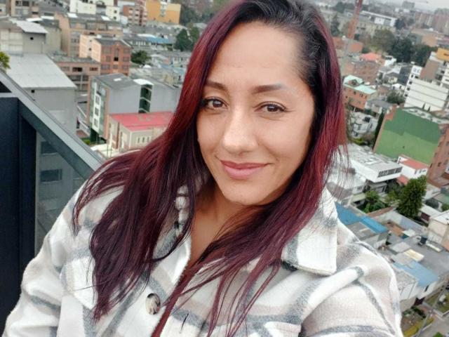Primer plano de SamanthaCorine, una mujer curvilínea con cabello largo y de color rojo oscuro, sonriendo ligeramente. Ella lleva una bata blanca y está de pie en un balcón con vistas a una bulliciosa ciudad. Muy atractivo/a..