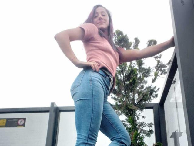 SamanthaCorine, de pie con confianza, con las manos en las caderas, vestida con una camiseta rosa ajustada y pantalones vaqueros azules, apoyada contra una edificio. Plano desde abajo que enfatiza su figura curvilínea y su actitud desafiante..