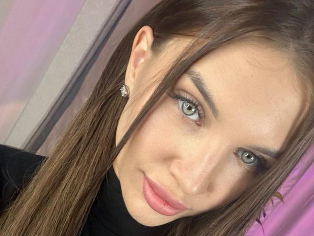 Gros plan de LunaRundlett, une jeune femme aux yeux verts clairs, aux longs cheveux bruns et aux lèvres roses. Elle porte un haut noir, des boucles d'oreilles en diamants et a un léger sourire. Extrêmement séduisant..