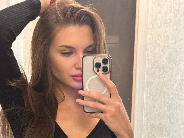 LunaRundlett, selfie, sosteniendo un iPhone con un agarre blanco, cabello castaño, labios rosados, blusa negra, brazo levantado, expresión seductora. mirada, sonrisa sutil, piel clara, luz natural, sombras suaves, informal, seguro de sí mismo, atractivo..