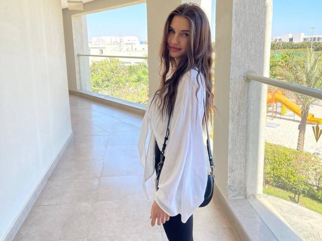 LunaRundlett with long, wavy brown hair, wearing a loose white shirt and black pants, stands in a sunlit corridor, looking over her shoulder. Bright sunlight highlights her face and hair.