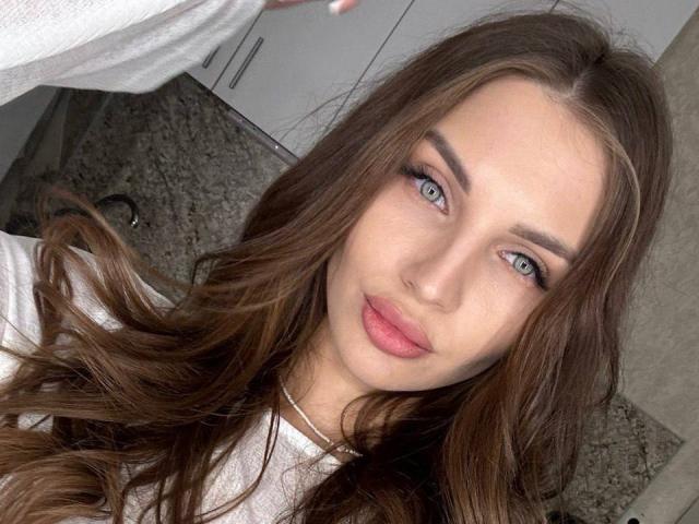 Primer plano de LunaRundlett con cabello castaño largo y ondulado, ojos azules y labios rosados. Ella lleva una blusa blanca y mira directamente a la cámara con una ligera sonrisa. Débiles sombras en su rostro.