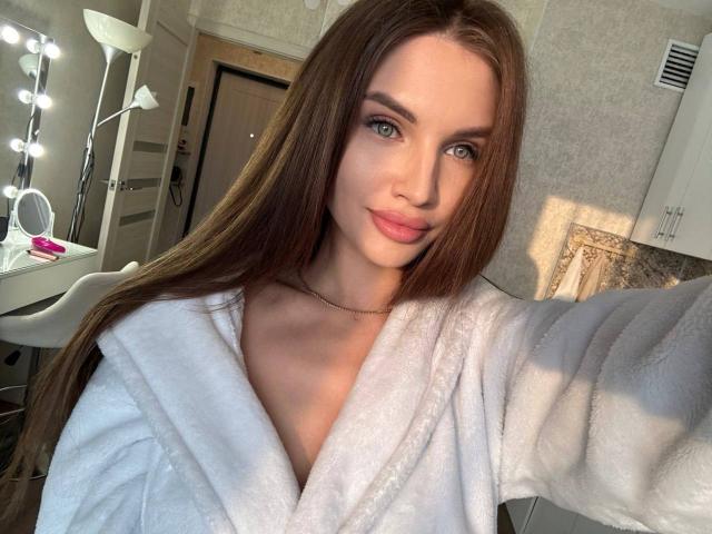 LunaRundlett, cabello castaño largo, ojos azules, bata blanca, luz solar suave, sonrisa sensual, selfie, brazo derecho extendido, lámpara. con bombillas redondas en el fondo, una ligera sombra en el rostro, ambiente casual e íntimo..