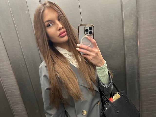 LunaRundlett, con lunghi capelli castani e un'espressione seria, sta scattando un selfie allo specchio con un iPhone, indossando una giacca grigia in stile militare. blazer con bottoni dorati, camicia verde, borsa nera, sfondo con ascensore, tensione sessuale.