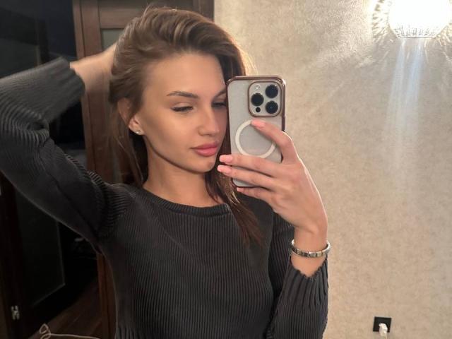 LunaRundlett, die ein Selfie mit einem silbernen iPhone macht, hat langes, braunes Haar und trägt einen schwarzen, gerippten Pullover. Sie sieht... Selbstbewusst, mit leicht geöffneten Lippen. Sie hält das Telefon mit einer Hand..