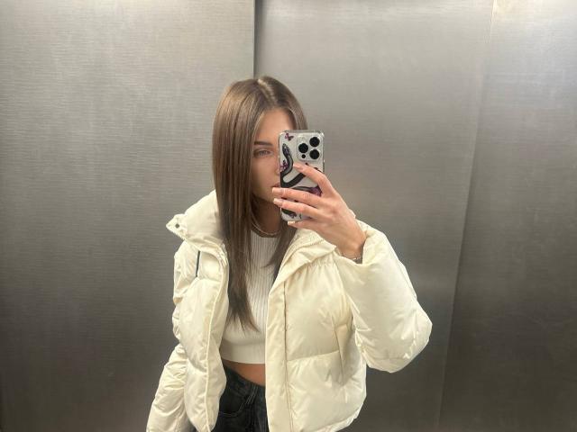 LunaRundlett, avec de longs cheveux bruns, prend un selfie devant un miroir, portant une veste matelassée blanche par-dessus un débardeur blanc, et tenant un objet noir. et téléphone blanc, sexy, décontracté, fond gris neutre.