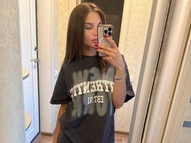 LunaRundlett está de pie en una habitación luminosa, tomando una foto de sí misma con un teléfono inteligente. Ella lleva una camiseta negra con letras blancas, tiene las uñas rojas y tiene el pelo largo y castaño. Muy atractivo..