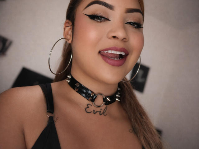 Primo piano di TatianaRossi, con i capelli castani raccolti in una coda di cavallo, che indossa una collana in pelle nera con la scritta "Evil" e un tatuaggio, e con un trucco marcato. eyeliner e rossetto viola, lingua di fuori, grandi orecchini a cerchio..