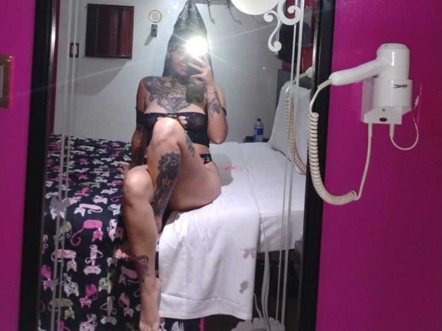 AmberKeller está sentada em uma cama, tirando uma selfie no espelho, usando lingerie preta, com tatuagens cobrindo os braços e as pernas, com uma perna levantada, expondo... coxa, clarão intenso que obscurece o rosto, lençol branco, parede rosa, secador de cabelo.