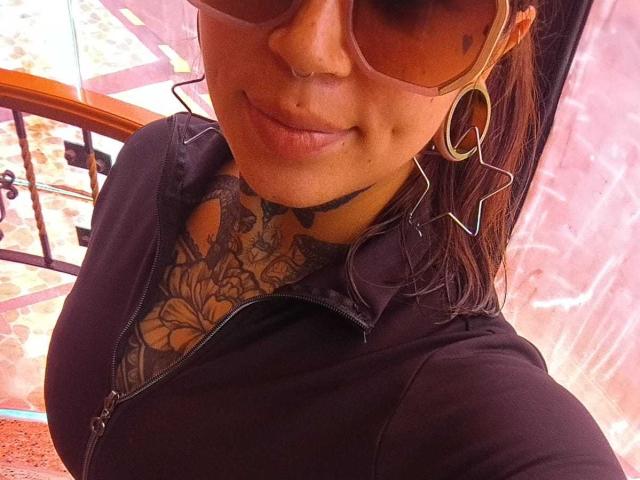 Primer plano de AmberKeller, mostrando su rostro y parte superior del cuerpo. Lleva grandes aros, gafas de sol y una sudadera con capucha negra, con un tatuaje floral visible en su pecho. Increíblemente sexy, con una ligera sonrisa..