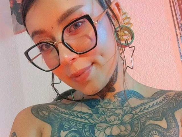 Photo en gros plan d'AmberKeller, portant des lunettes à monture noire, avec des cheveux foncés attachés en chignon, de grandes boucles d'oreilles vertes, et tatouage floral bleu détaillé sur toute la poitrine. Fond rose clair. Incroyablement sexy..