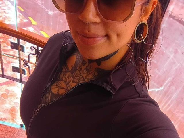 Close-up photo of AmberKeller with tan skin, wearing large sunglasses, black top revealing intricate chest tattoo, hoop earrings, and black choker, leaning slightly forward with a subtle smile. Background includes wrought iron railing.