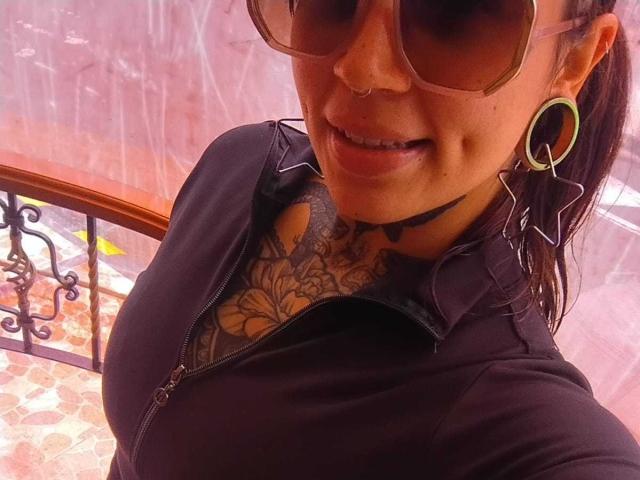 Close-up photo of AmberKeller, a tattooed woman with large breasts, wearing dark sunglasses, black zip-up shirt, and green hoop earrings, showing a seductive smile, with visible sexual tension.