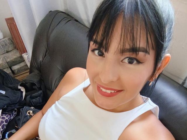 Selfie de primer plano de TalianaSquirt con cabello oscuro, flequillo y delineador de ojos negro, sonriendo con labios rojos. Ella lleva una camiseta blanca y está recostada en un sofá de cuero negro. Su pene no es visible..