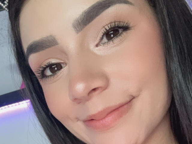 Gros plan de KimmyK avec des cheveux foncés, des sourcils épais et des yeux marron. Elle sourit discrètement, avec de belles lèvres pulpeuses et une peau naturelle. La lumière illumine son visage, créant un éclat doux. Légère tension sexuelle.