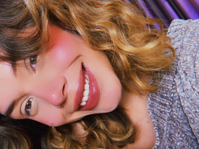 Primer plano de EmmaRiveraa, sonriendo con un ligero rubor rosado en las mejillas, cabello castaño rizado y dientes blancos. Ella lleva una blusa gris con textura. Su expresión es sensual y atractiva..